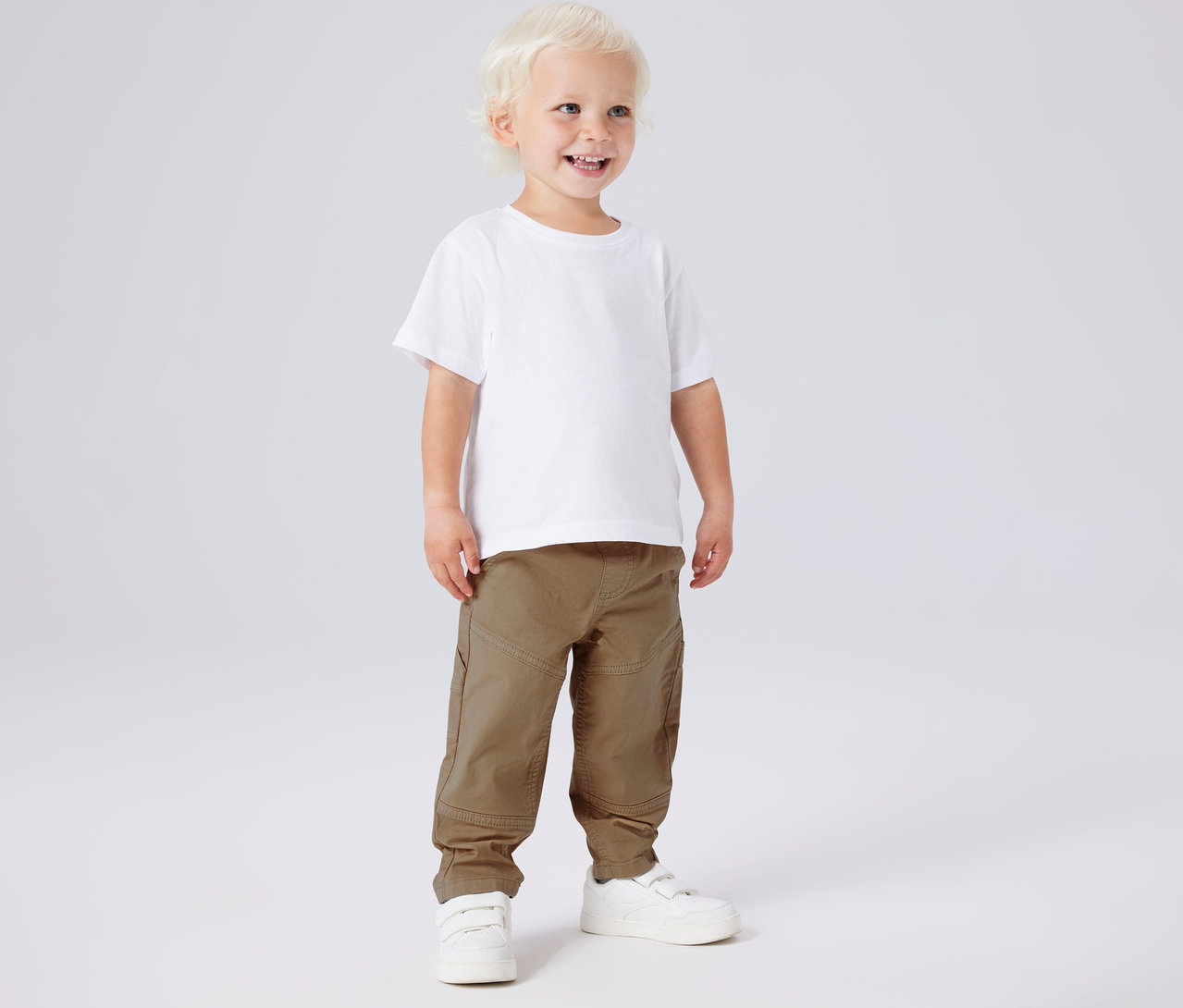 Lächelnder Junge mit weißem T-Shirt und NAME IT Kinder-Twillhose in Khaki und weißen Schuhen.
