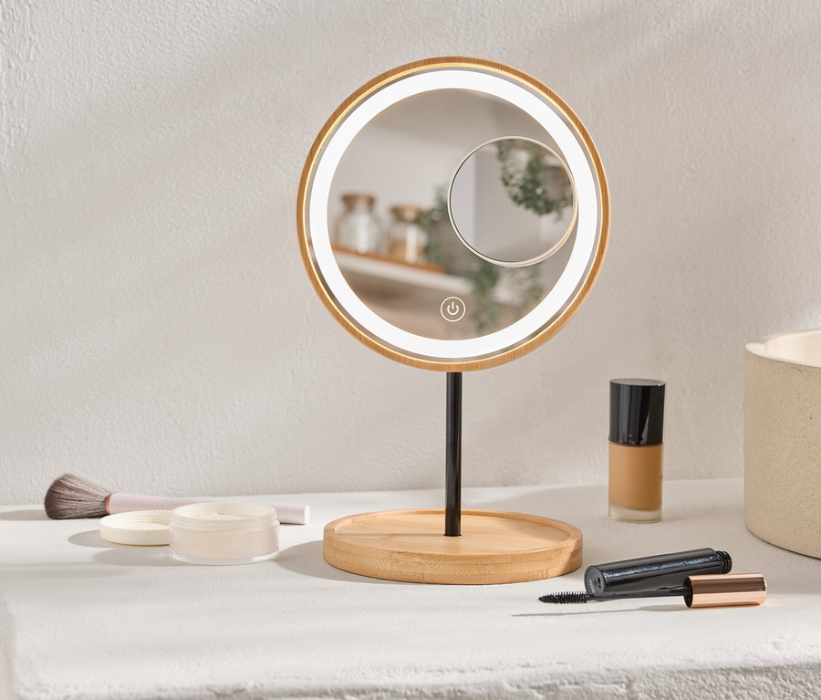 Auf einer Ablage stehen ein runder, beleuchteter Schminkspiegel, Make-up und ein Pinsel.