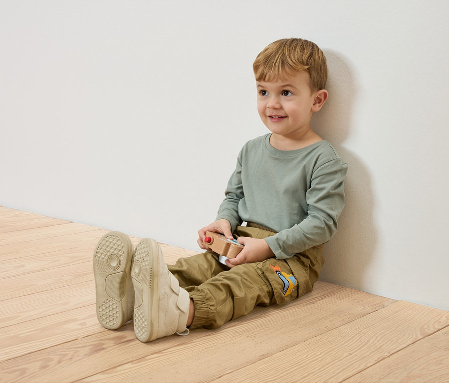 Ein lächelnder Junge sitzt auf dem Holzboden, an eine weiße Wand gelehnt, und hält ein hölzernes Spielzeugauto.