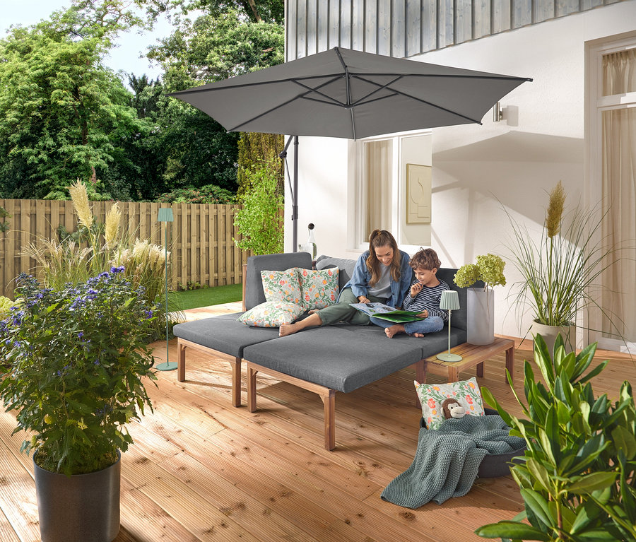 Eine Frau und ein Kind sitzen auf einem grauen Gartensofa mit separat ausziehbaren Fußstützen. Auf dem Sofa liegen zwei Outdoor-Kissen mit floralem Muster. Ein anthrazitfarbener Leco Ampelschirm mit Schirmständer und Abdeckhaube spendet Schatten. Eine salbeifarbene LED-Akku-Outdoor-Tischleuchte steht auf einem Holztisch neben dem Sofa, während eine salbeifarbene LED-Akku-Outdoor-Stehleuchte im Hintergrund zu sehen ist.