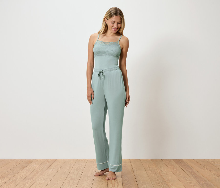 Frau trägt einen hellgrünen Schlafanzug mit Spitzendetails, stehend auf einem Holzboden vor einer weißen Wand.