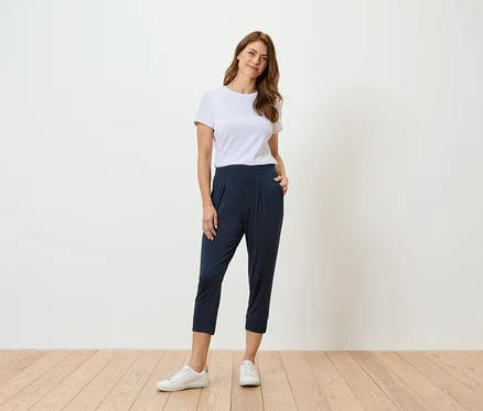 Frau in weißem T-Shirt und marineblauer Hose steht vor einer weißen Wand.