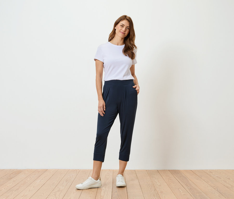 Frau in weißem T-Shirt und marineblauer Hose steht vor einer weißen Wand.