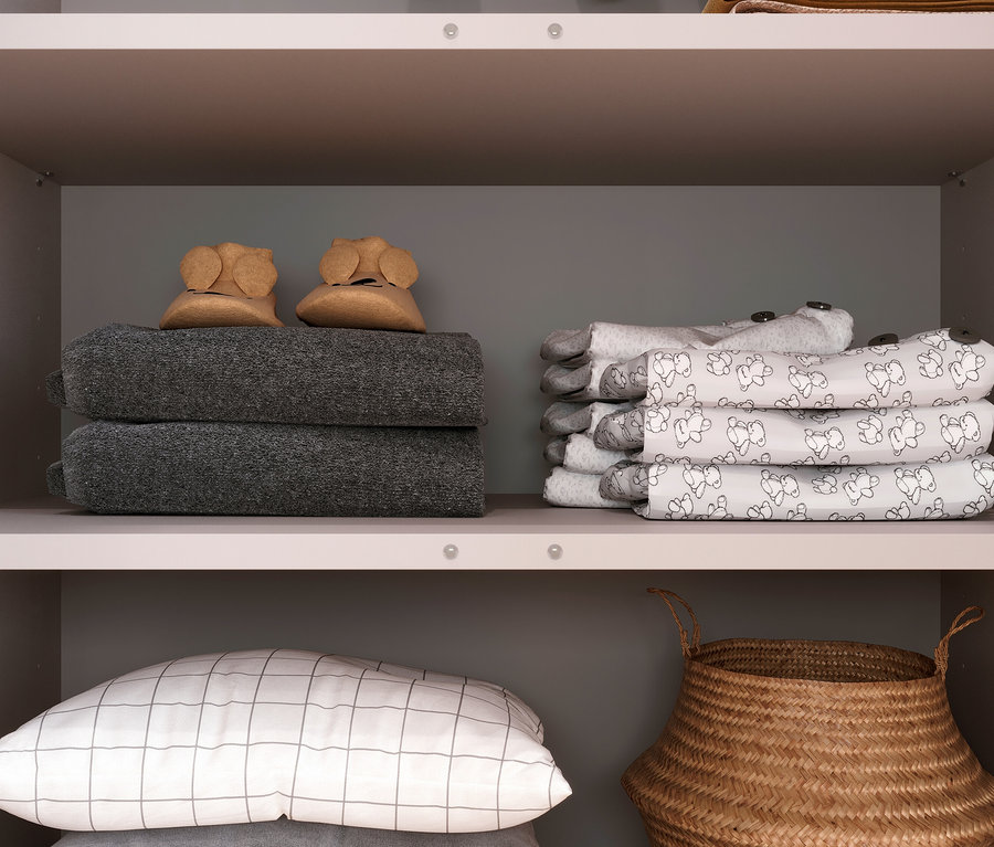 Aufnahme eines Schranks mit grauem Hintergrund, in dem sich zusammengelegte graue Handtücher, Babyschuhe, Stofftiere, Kissen und ein Korb befinden.
