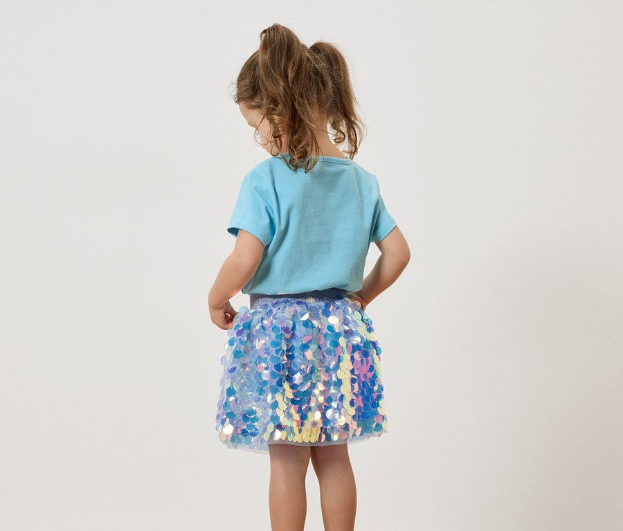 Ein junges Mädchen mit Zöpfen trägt ein hellblaues T-Shirt und einen glitzernden Paillettenrock.
