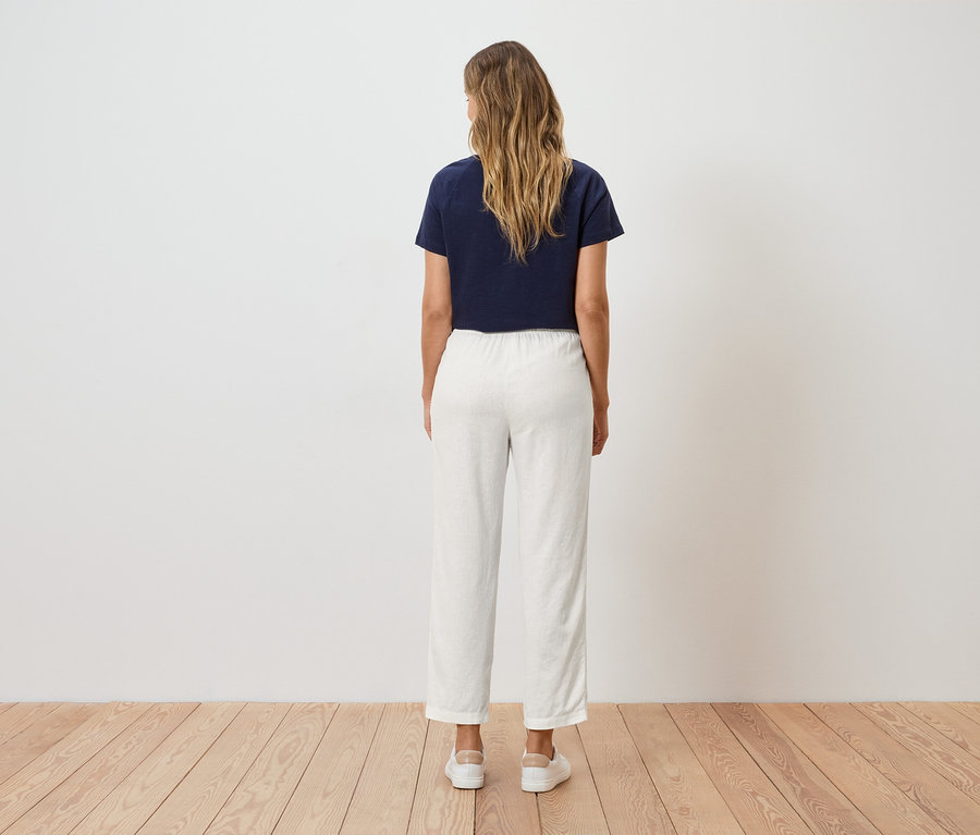 Eine Frau mit blonden, gewellten Haaren steht mit dem Rücken zum Betrachter, trägt ein marineblaues T-Shirt und eine weiße Hose.