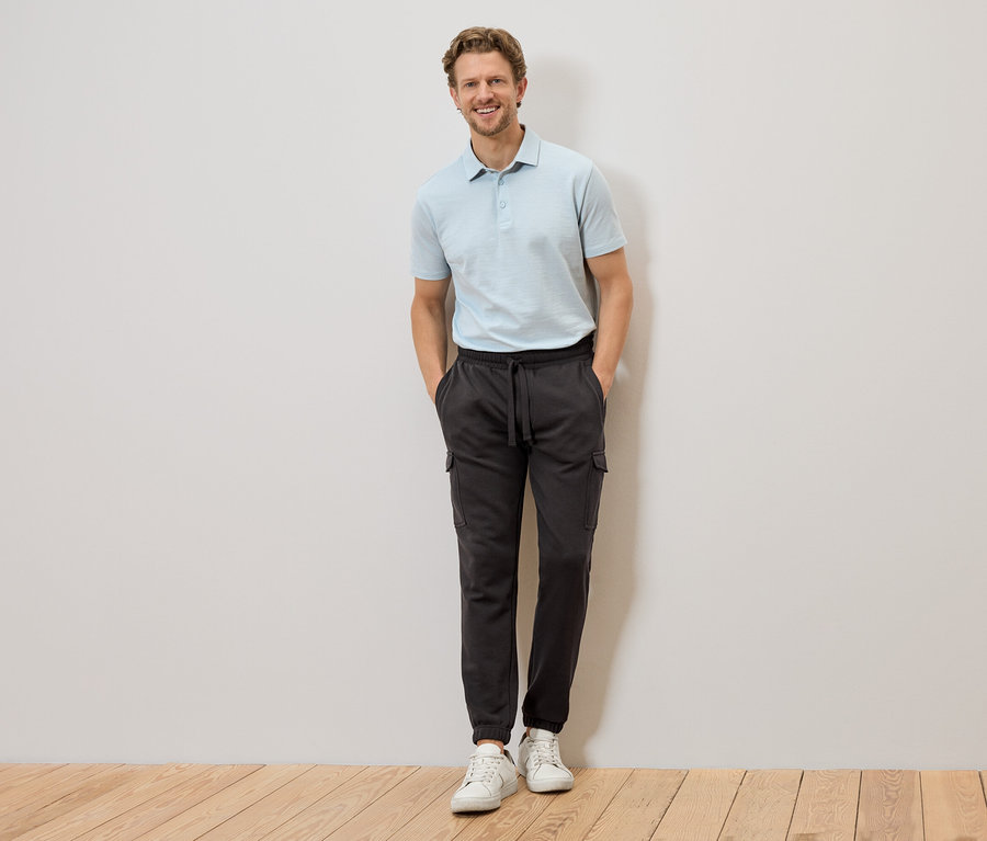Ein Mann mit hellblauem Poloshirt und grauer Cargohose steht mit den Händen in den Hosentaschen vor einer Wand.