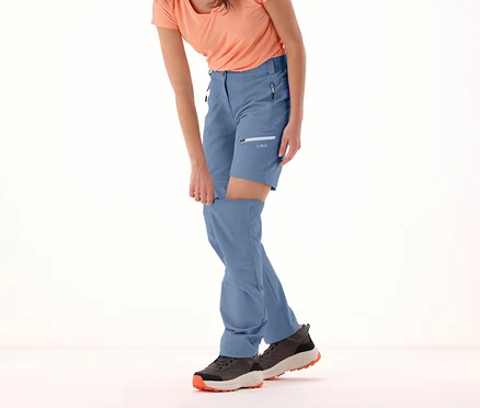 Frau, die die Beine der blauen CMP Damen-Zip-off-Hose abzippt. Sie trägt dazu ein T-Shirt und Wanderschuhe.