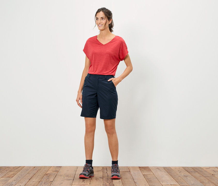 Frau steht mit der Hand in der Hosentasche, trägt ein rotes T-Shirt und blaue Shorts.