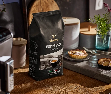 Eine Packung Tchibo Espresso Kräftig Ganze Bohne steht auf einem Holztisch neben einer Kaffeemaschine und Tellern mit Törtchen.
