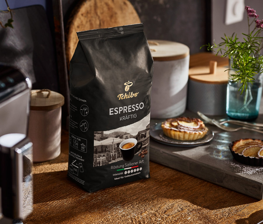 Eine Packung Tchibo Espresso Kräftig Ganze Bohne steht auf einem Holztisch neben einer Kaffeemaschine und Tellern mit Törtchen.