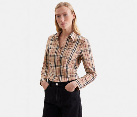 Blonde Frau trägt ein beiges, langärmeliges Karoshirt mit schwarzen, roten und weißen Linien und schwarze Jeans mit den Händen in den Taschen, vor weißem Hintergrund stehend.