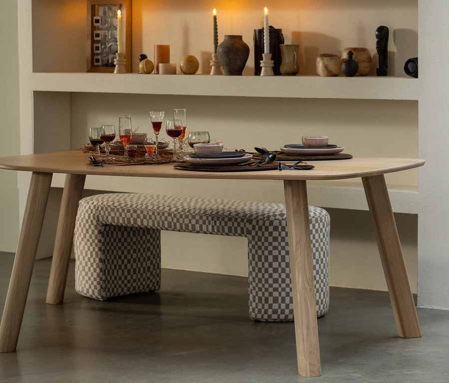 Naturfarbener WOOOD Tisch »Tablo« (ca. 180 x 100 cm) mit Gedeck für ein Abendessen. Darunter eine karierte Bank. Im Hintergrund Kerzen und Dekorationen auf einem Regal.