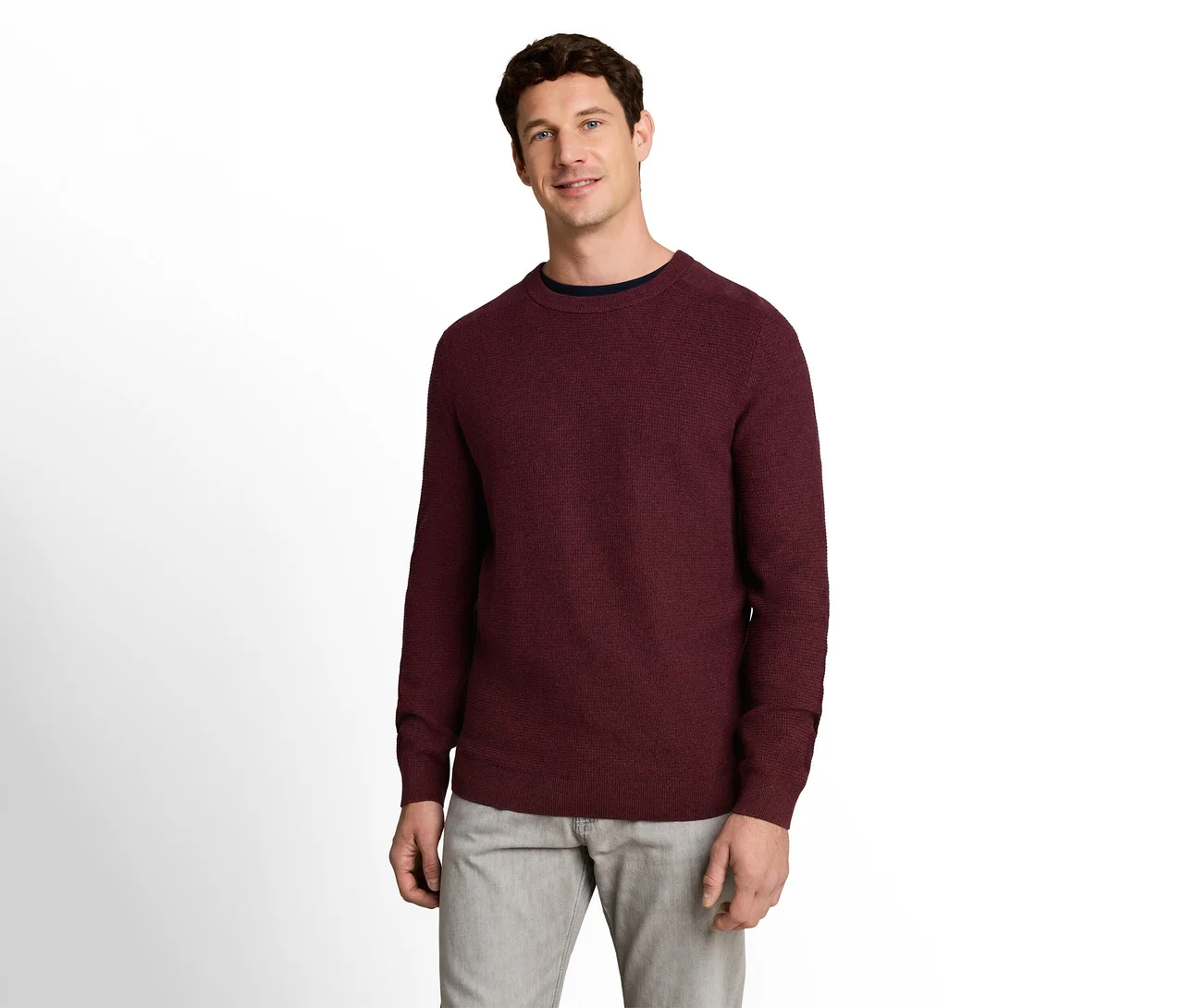 Mann in einem weinroten Pullover mit Rundhalsausschnitt über einem dunkelblauen T-Shirt und hellgrauen Hosen, lächelnd.