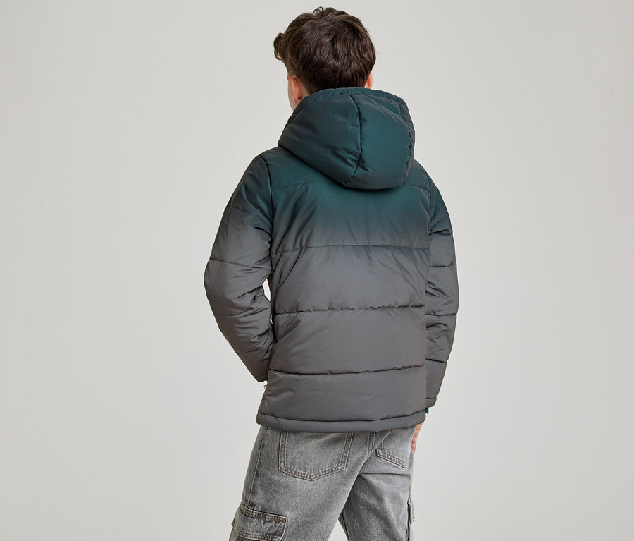 Ein junger Mann von hinten, trägt eine Kapuzen-Steppjacke mit Farbverlauf von Dunkeltürkis zu Dunkelgrau und graue Jeans. Seine Hände sind in den Taschen.