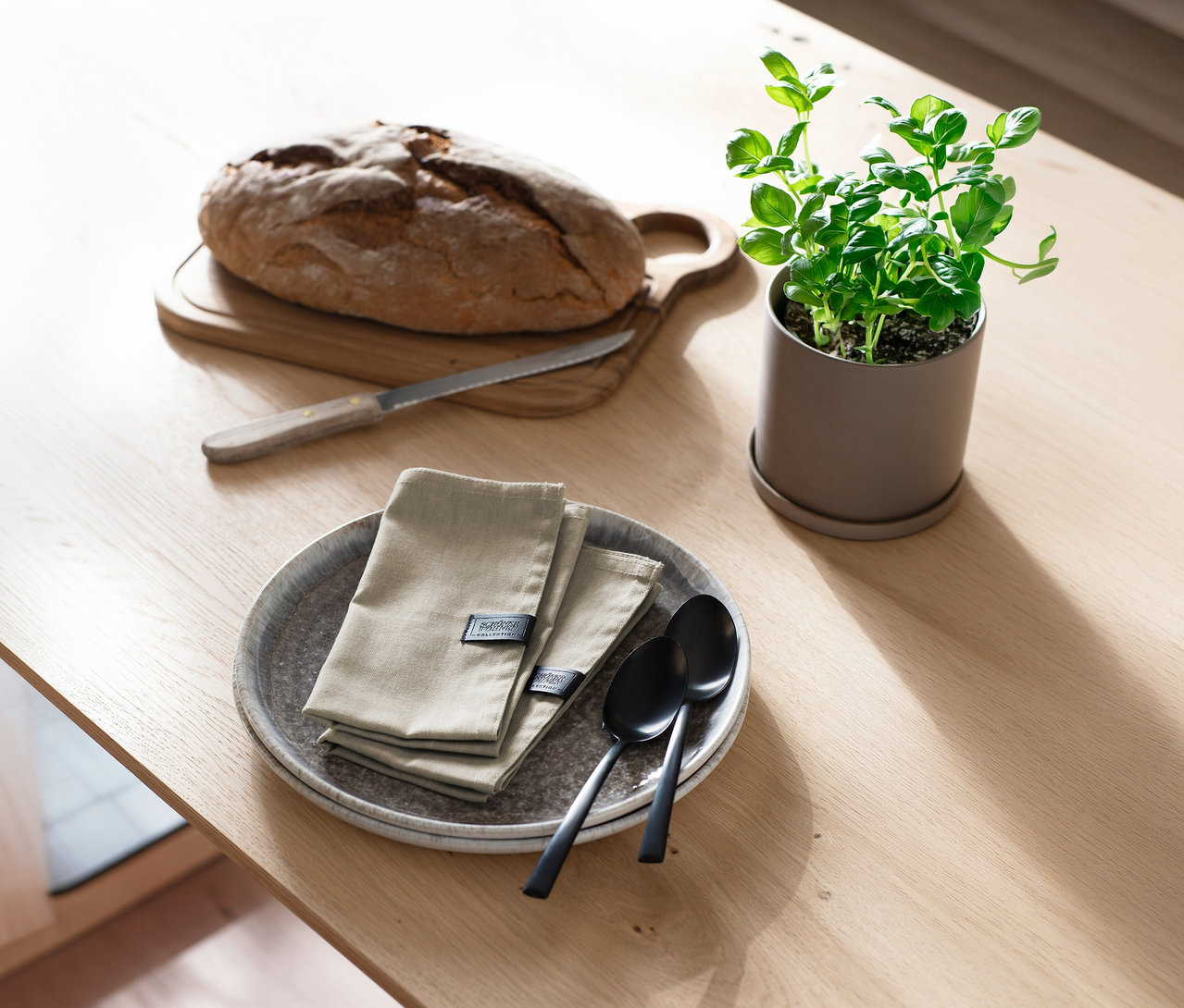 Ein Laib Brot auf einem Holzschneidebrett mit Brotmesser, daneben zwei sandfarbene SCHÖNER WOHNEN-Kollektion Servietten auf grauen Tellern mit zwei schwarzen Löffeln und eine Topfpflanze auf einem Holztisch.