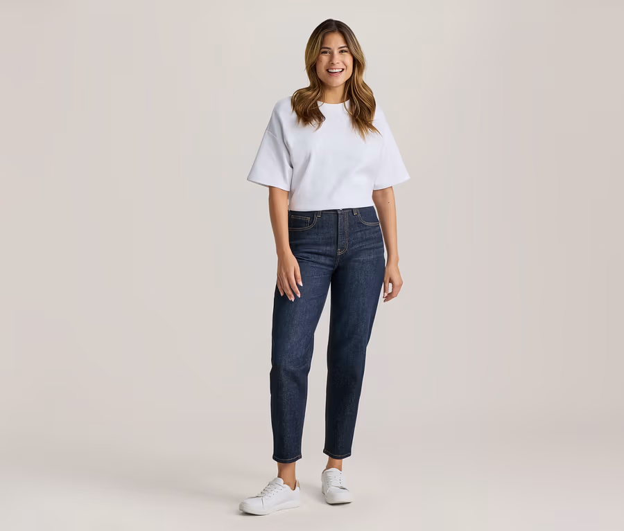 Frau steht lächelnd in weißem T-Shirt, dunkler Jeans und weißen Turnschuhen.