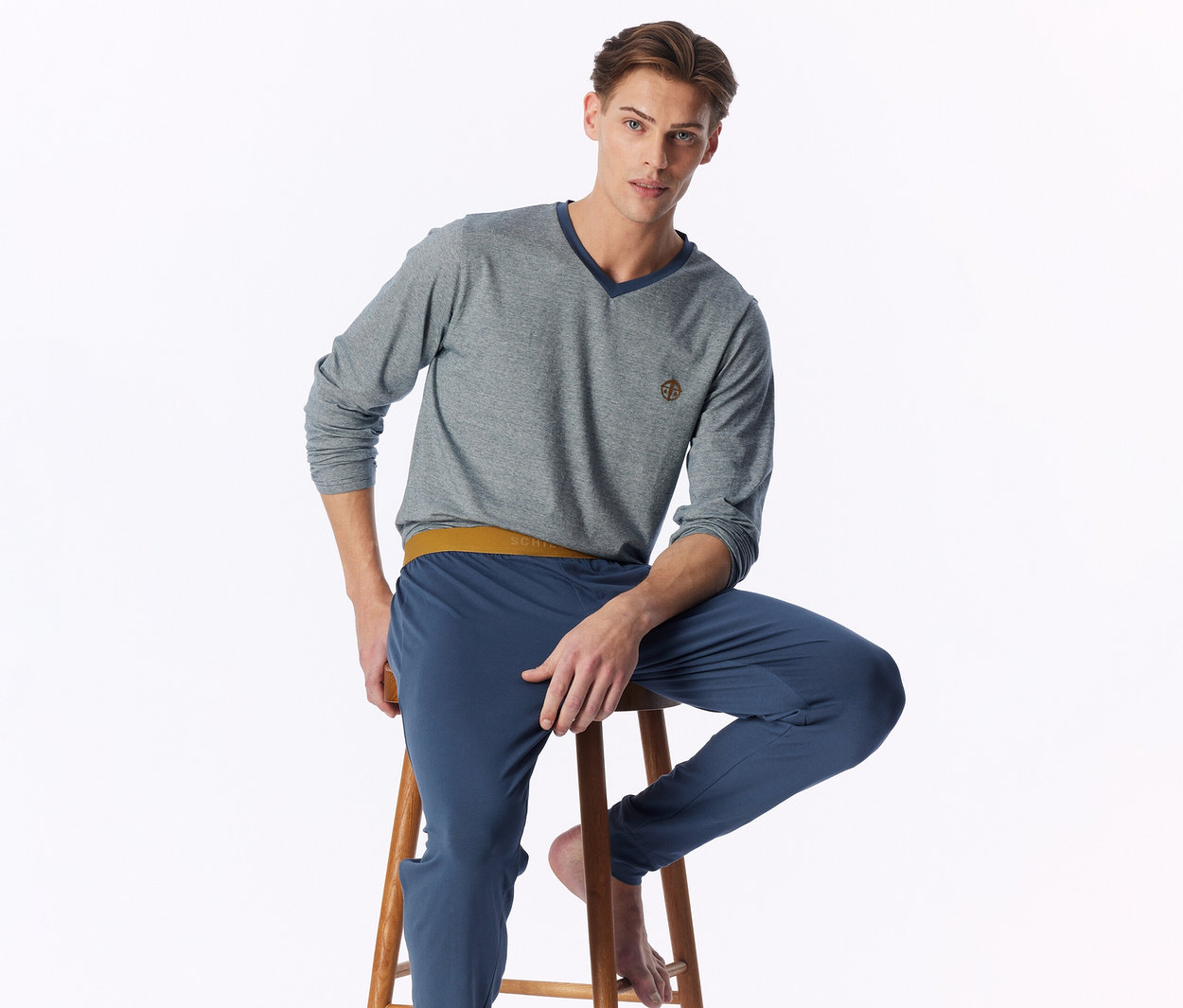 Ein Mann sitzt auf einem Hocker, blickt in die Kamera und trägt ein graues Langarmshirt mit V-Ausschnitt und Emblem sowie blaue Jogginghosen mit senfgelbem Bund. Er hat nackte Füße.