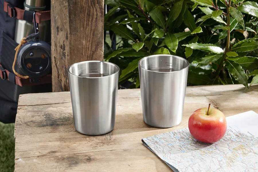 Zwei Camping-Becher aus Edelstahl stehen neben einem Apfel und einer Landkarte auf einem Holztisch.