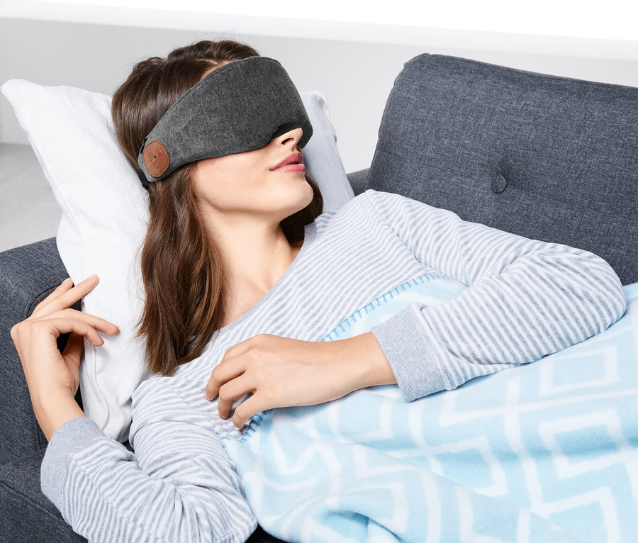 Frau liegt mit Schlafmaske mit Bluetooth®-Kopfhörern auf Couch.