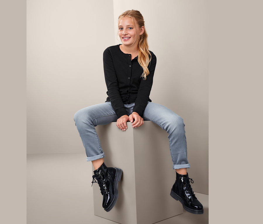 Mädchen sitzt auf einem Würfel und trägt eine Feinstrickjacke, Jeans und schwarze Stiefel.