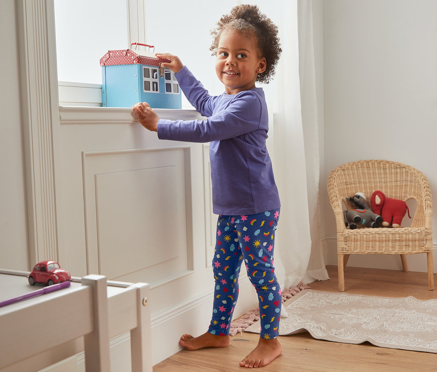 Mädchen steht am Fenster und spielt mit Spielzeughaus, trägt 5 Jersey-Leggings aus Bio-Baumwolle.
