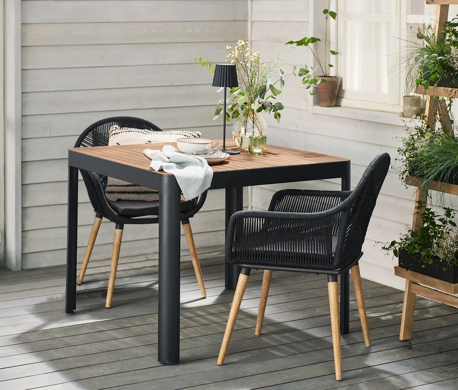 Outdoor-Terrasse mit einem quadratischen Gartentisch mit abgerundeten Beinen und zwei Schalenstühlen mit Textilgeflecht. Auf dem Tisch stehen eine LED-Akku-Tischleuchte und Blumen. Rechts daneben eine zusammenklappbare Pflanzleiter mit schwarzen Pflanzkästen voller Grünpflanzen.