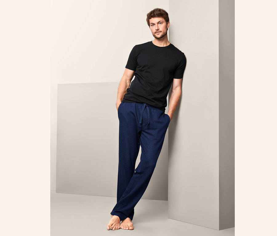 Mann lehnt an einer Wand, trägt ein schwarzes T-Shirt und blaue Hosen. Er trägt 2 T-Shirts mit Rundhals-Ausschnitt.