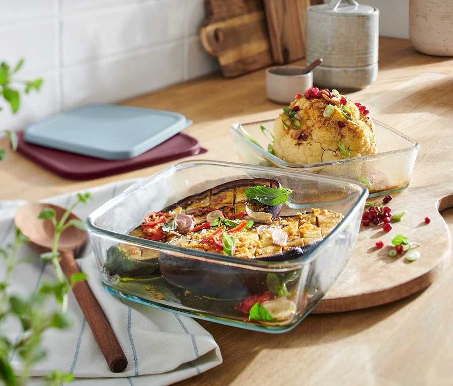 Zwei Glas-Auflaufformen mit Deckel, eine mit Aubergine und Zucchini, die andere mit Blumenkohl, auf einem Holzbrett.