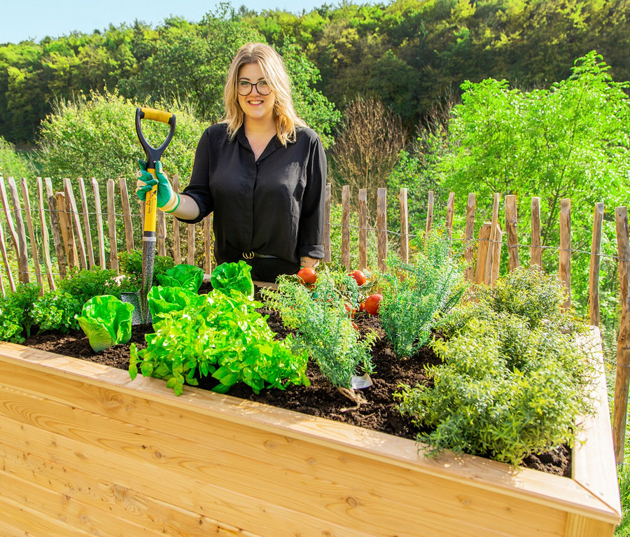 Frau steht hinter einem 170 cm großen Hochbeet aus Lärche und hält einen Spaten.