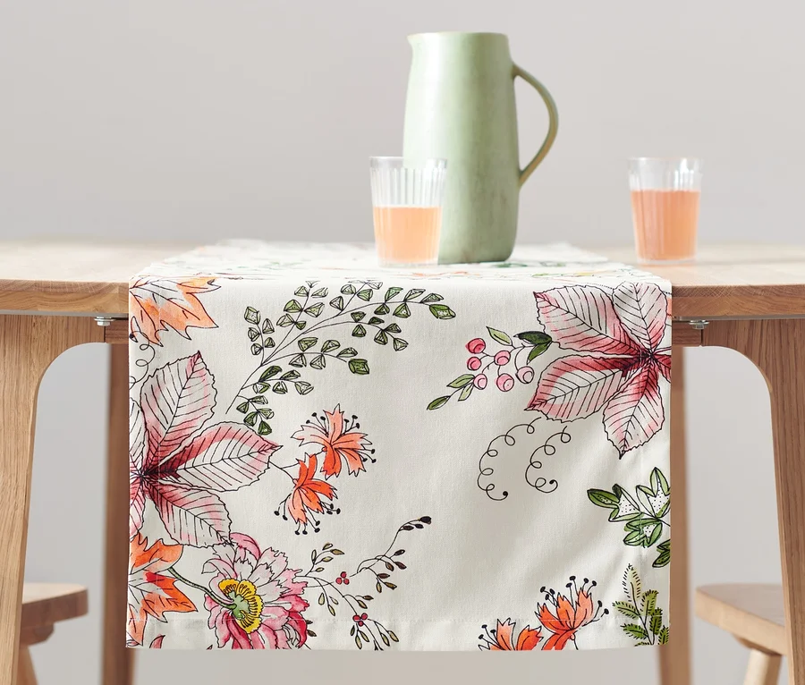 Auf einem Holztisch liegt ein Tischläufer mit Blumenmuster. Auf dem Tisch stehen ein Krug und zwei Gläser mit orangefarbener Flüssigkeit.