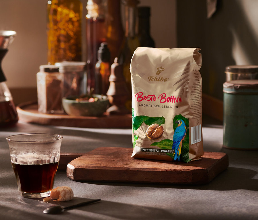 Eine Packung Beste Bohne - 500 g Ganze Bohne steht auf einem Holzbrett neben einem Glas Kaffee.