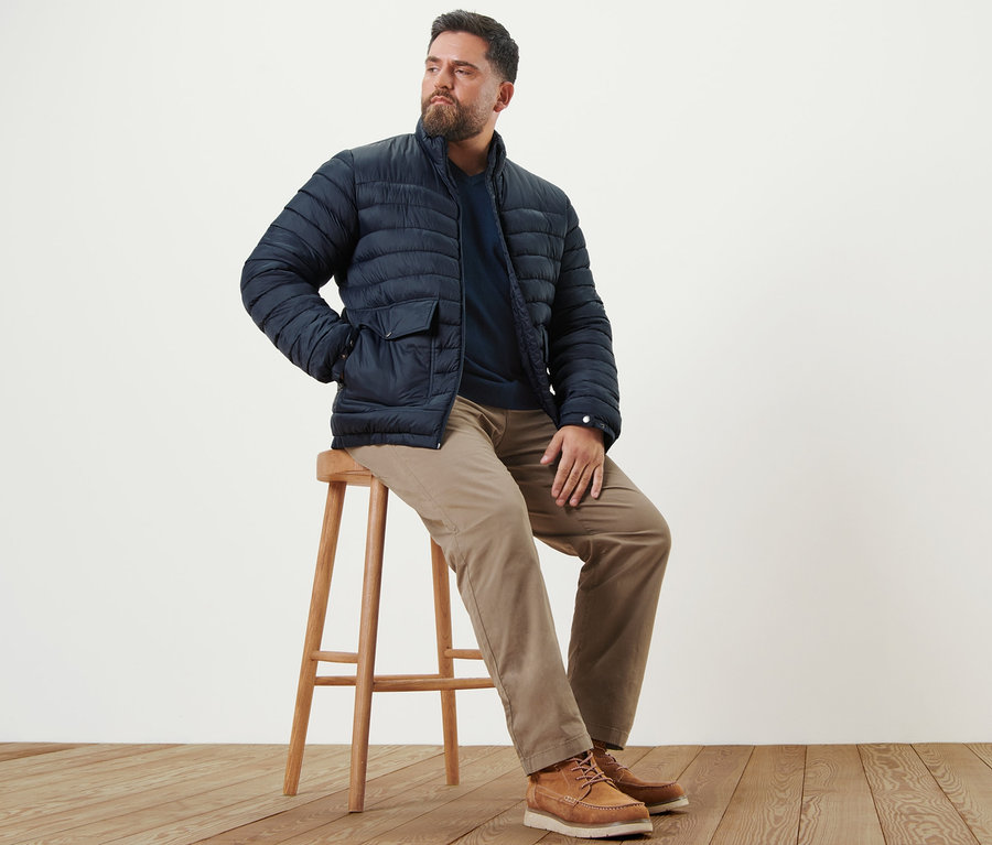 Mann sitzt auf einem Hocker und trägt sandfarbene Chino, einen marineblauen Pullover mit V-Ausschnitt und eine marineblaue Steppjacke.