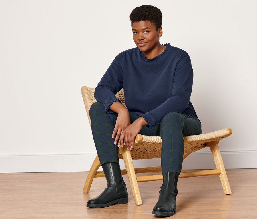 Frau sitzt auf einem Korbstuhl und trägt eine Stretchhose, grün-blau kariert, Lederboots und ein Sweatshirt mit Strickdetails.