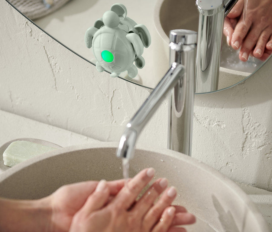 Eine Person wäscht sich die Hände in einem Waschbecken, an einem Spiegel ist ein Händewasch-und-Zahnputz-Timer angebracht.