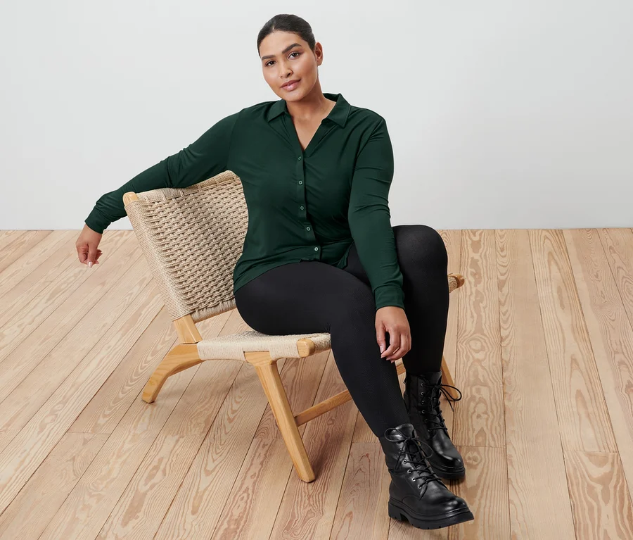 Eine Frau sitzt auf einem Korbstuhl und trägt ein dunkelgrünes Blusenshirt mit Knopfleiste, Thermo-Leggings mit Prägung und Leder-Boots.