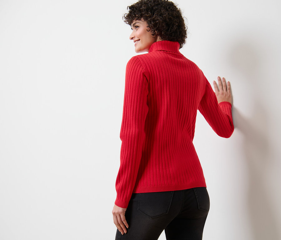 Frau lehnt an einer weißen Wand und trägt einen gerippten Rollkragenpullover in Rot.
