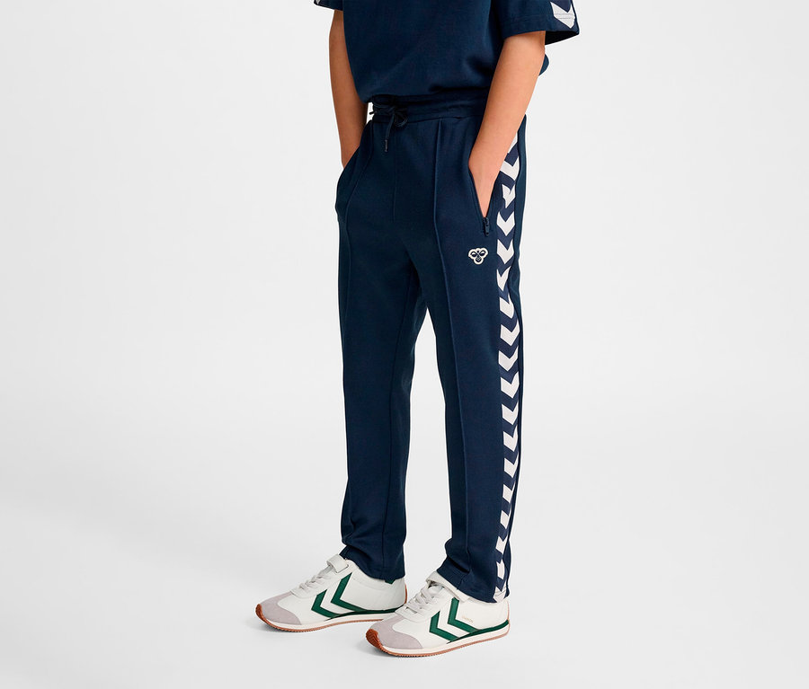 Ein Junge steht und trägt eine marineblaue HUMMEL HMLJR Archive Regular Poly Pants und Turnschuhe.