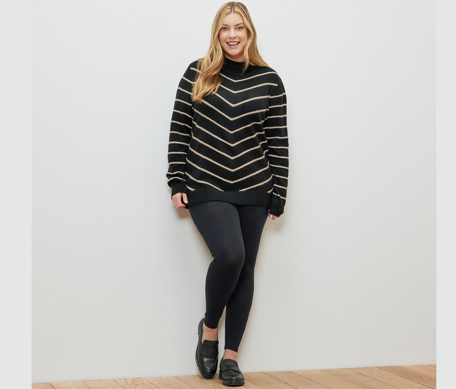 Frau posiert in einem schwarz gestreiften Strickpullover mit Stehkragen und Leggings.
