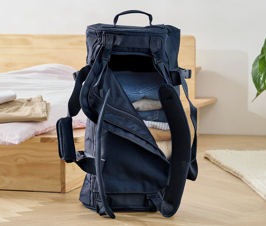 Eine offene Reisetasche-und-Rucksack, gefüllt mit Kleidung, steht auf dem Boden neben einem Bett.