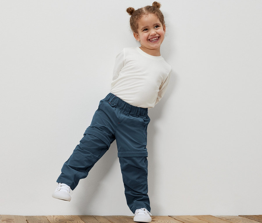 Lächelndes kleines Mädchen, das sich an eine weiße Wand lehnt und eine Kleinkinder-Funktionshose und ein weißes Hemd trägt.