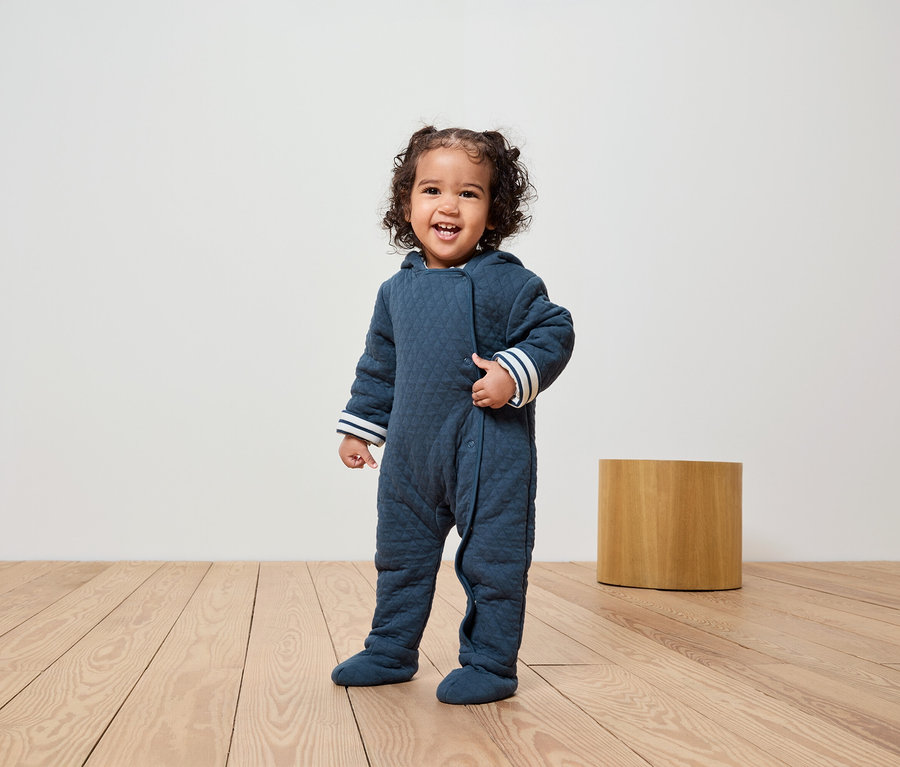 Ein lächelndes Kind steht auf einem Holzboden und trägt einen marineblauen, gesteppten Overall mit Kapuze und weißen Streifen an den Bündchen.