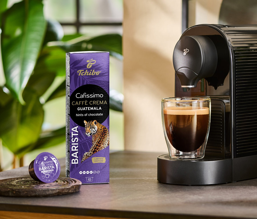 Kaffeemaschine bereitet Cafissimo BARISTA Caffè Crema Guatemala in einem Glas zu. Daneben stehen eine 10-Kapsel-Packung und eine Kapsel.