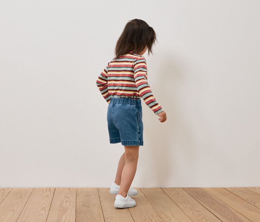 Mädchen mit gestreiftem Langarmshirt und Jeansshorts steht mit dem Rücken zur Kamera.