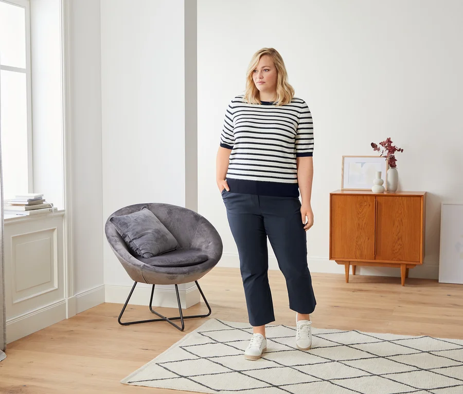Eine Frau steht in einem Raum auf einem Teppich und trägt eine Cropped Bengalin-Hose, Canvas-Sneaker und einen Feinstrickpullover mit Halbarm.