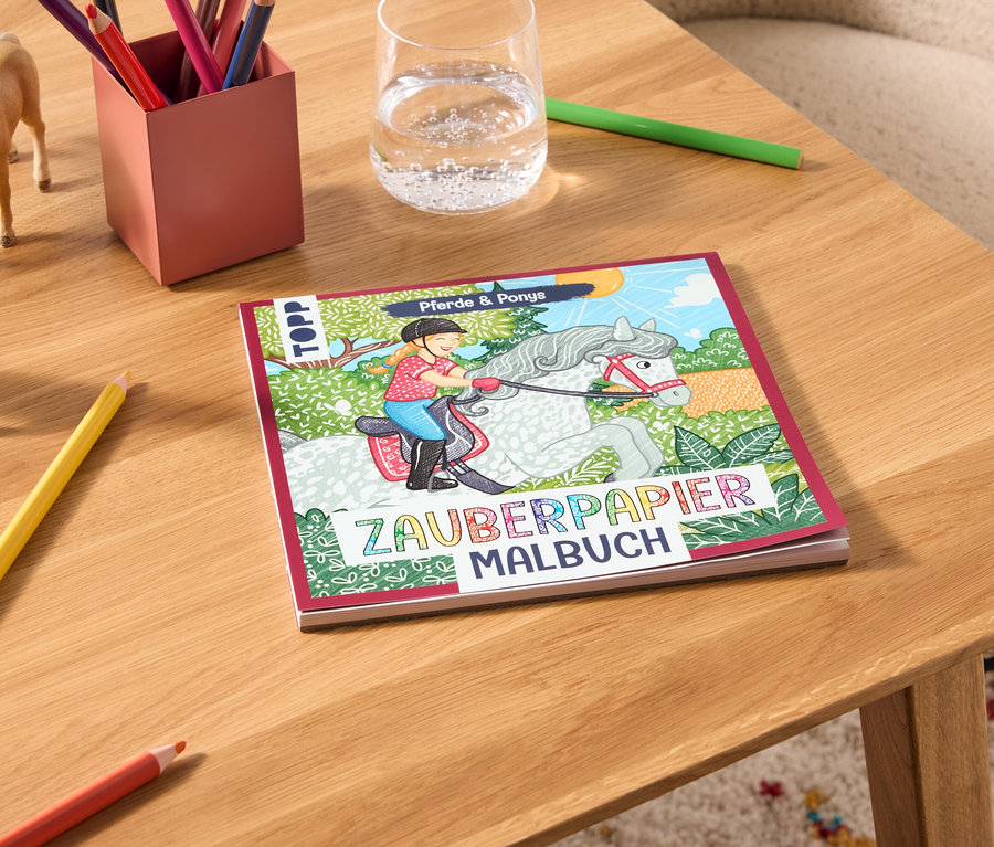 Auf einem Tisch liegen ein Zauberpapier Malbuch, Buntstifte, ein Glas Wasser und zwei Pferdefiguren.