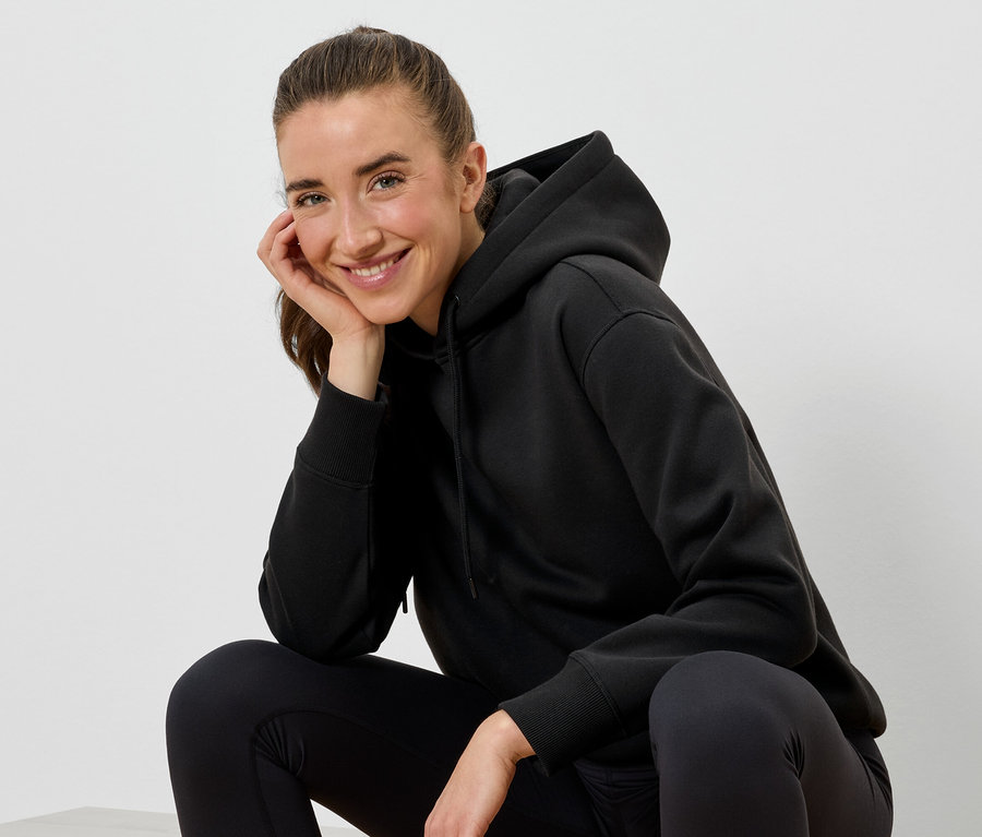 Eine lächelnde Frau in einem schwarzen Hoodie und schwarzen Leggings sitzt und stützt ihr Gesicht mit der Hand.