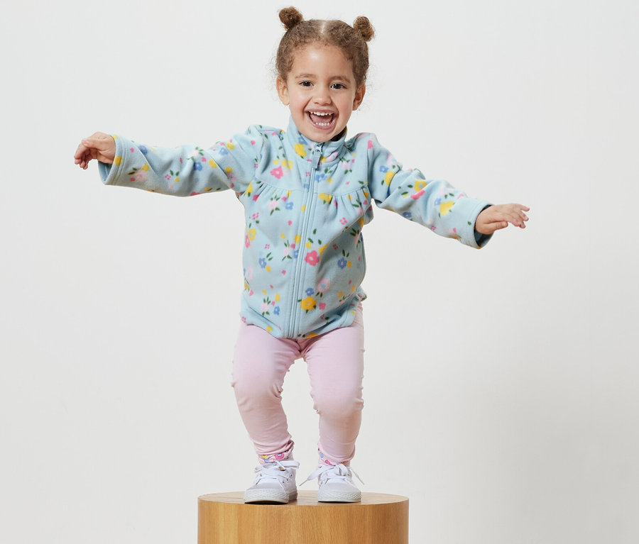 Lächelndes kleines Mädchen steht auf einem Holzsockel und trägt eine hellblaue Fleecejacke.