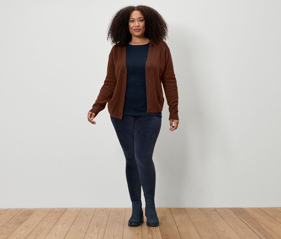 Eine Frau mit braunem lockigem Haar steht vor einer weißen Wand und trägt eine braune Strickjacke, ein marineblaues T-Shirt, marineblaue Leggings und marineblaue Stiefel.