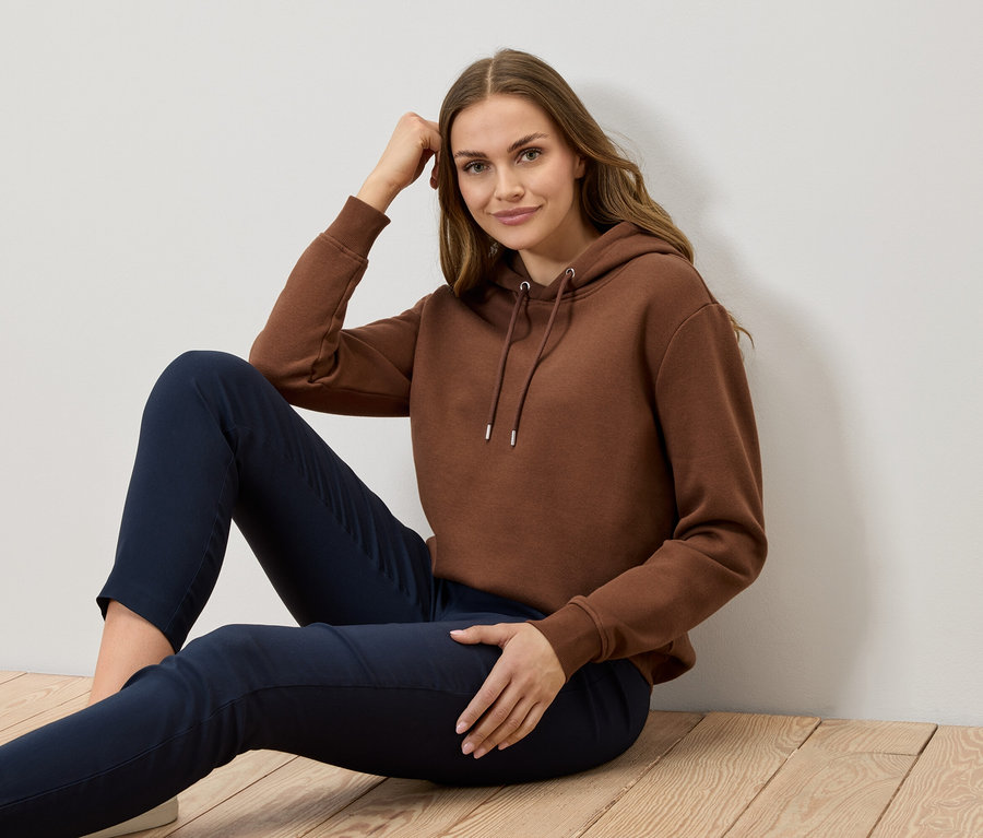 Eine Frau sitzt auf einem Holzboden und trägt einen braunen Hoodie und dunkelblaue Hosen.